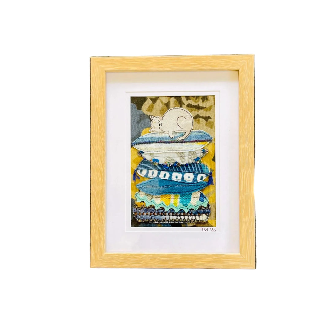 Framed textile art featuring a whimsical illustration of a white cat lounging on a blue and yellow striped pedestal, set in a light natural wood frame with a white mat against a white wall.