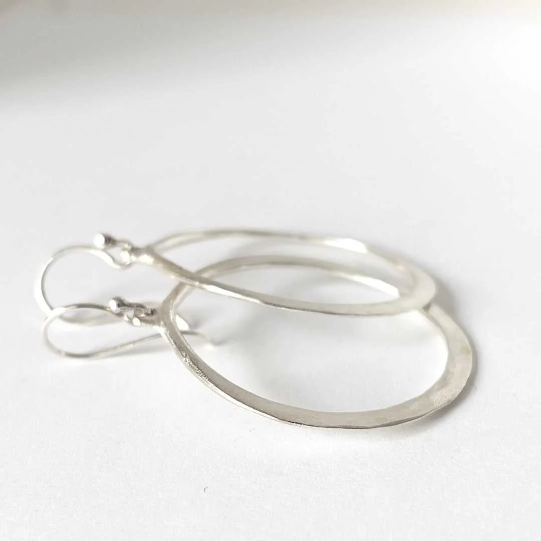 A pair of minimalist silver hoop earrings with textured, hammered finishes and matching fishhook ear wires, displayed on a clean white surface.