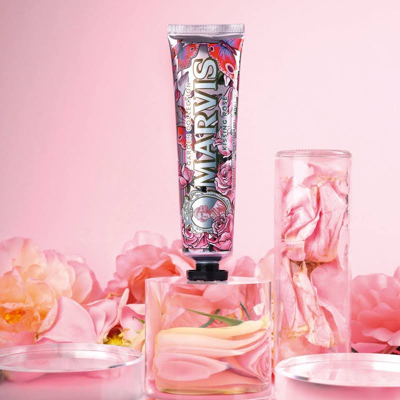 A squeeze tube of Marvis Kissing Rose toothpaste stands upright on a clear glass base, surrounded by pink rose petals and dried rose petals in glass vessels against a soft pink background.