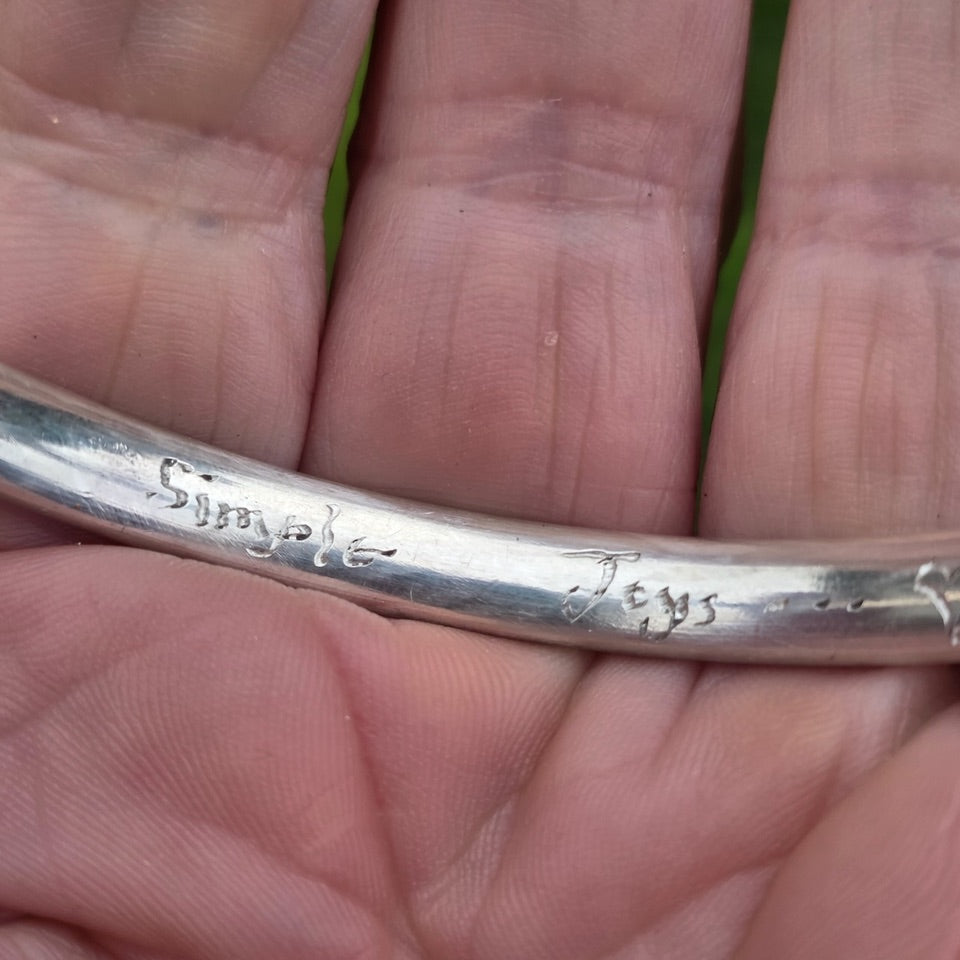 A close-up of a silver bangle held in a person's hand, showing engraved script markings on the polished surface.