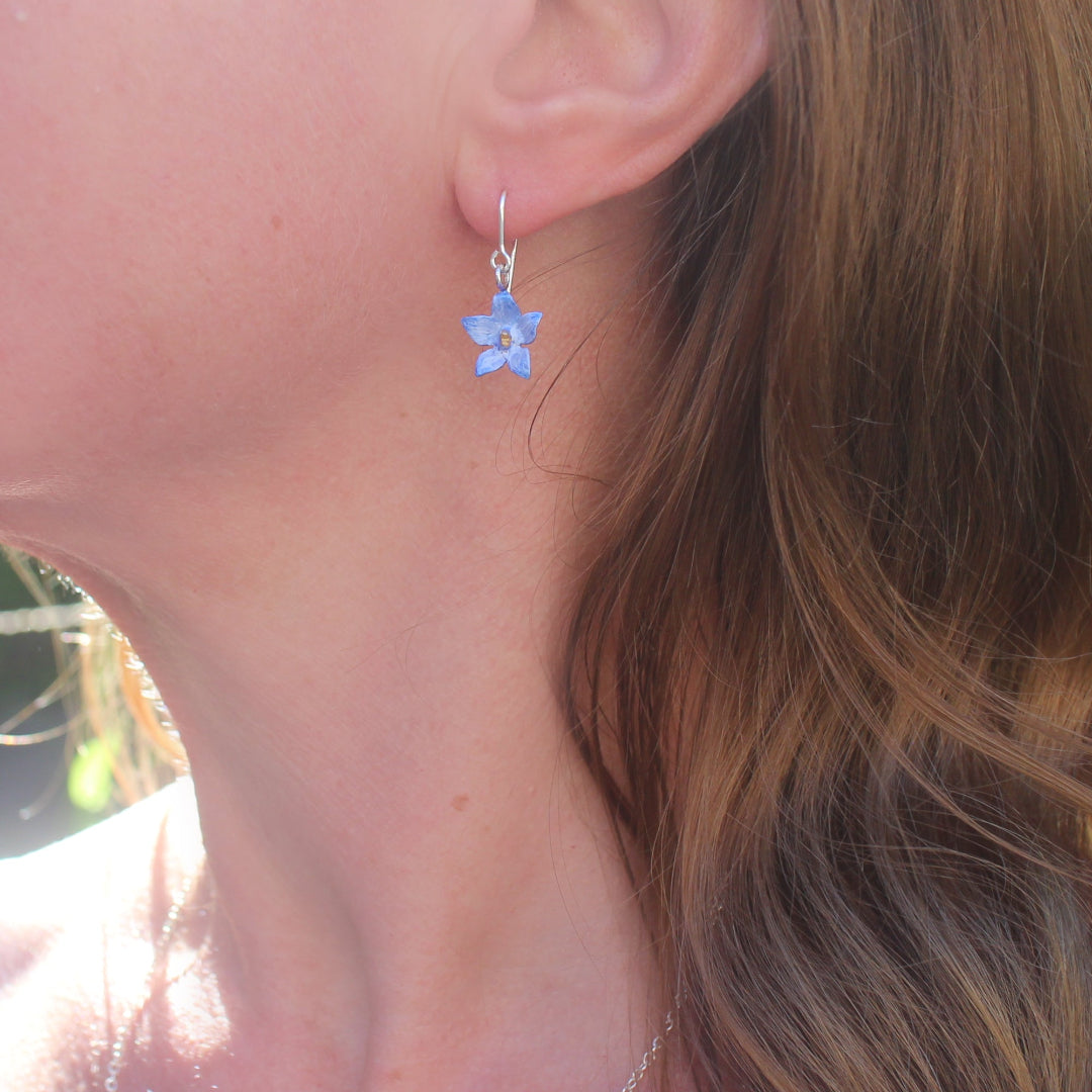 A single blue and purple star-shaped sterling silver earring with a floral design, hanging from a sterling silver ear hooks on a persons ear.