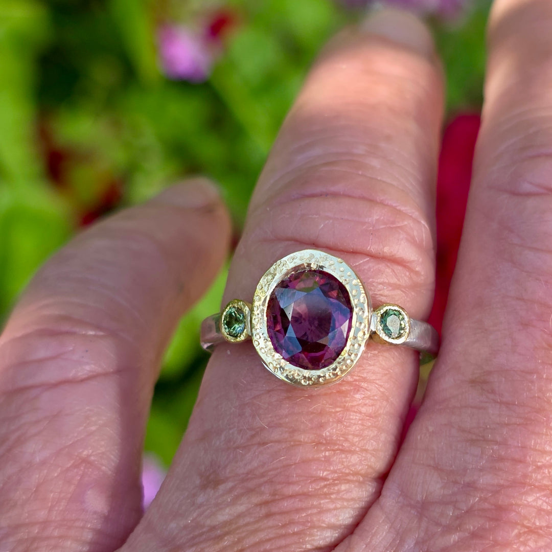 Ring with a red zircon gemstone & green sapphires on a finger against a blurred natural background