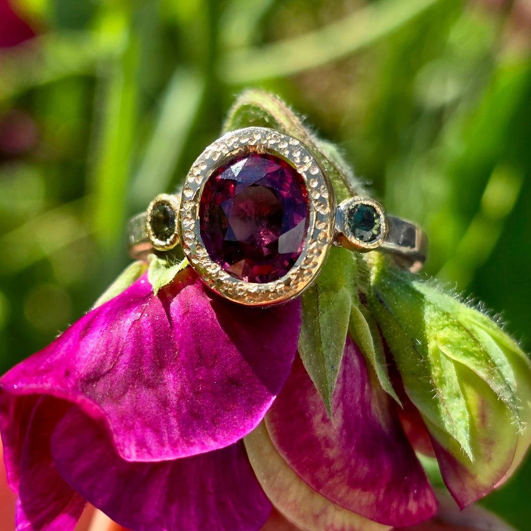 Ring with a red zircon gemstone & green sapphires on a pink flower with a green blurred background