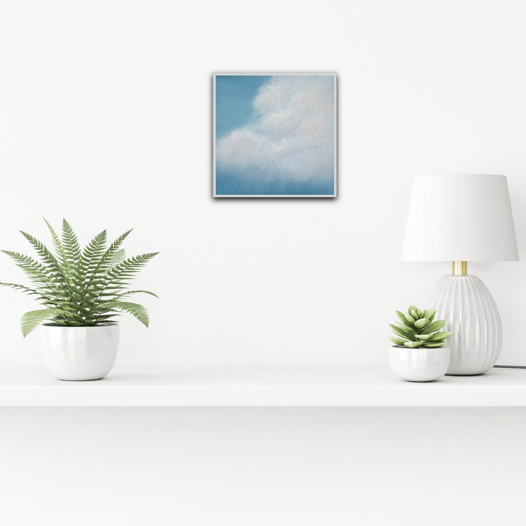 Fern plant, lamp, and small plant on a white surface with a framed cloud art print above.