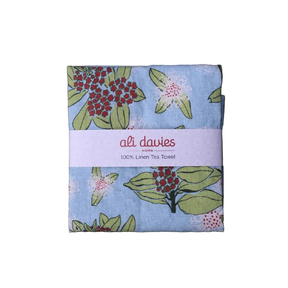 A folded linen tea towel with a botanical pattern of red berries and green leaves on a light blue background, wrapped in a white paper band that reads 'ali dawies 100% Linen Tea Towel', set against a white  background.