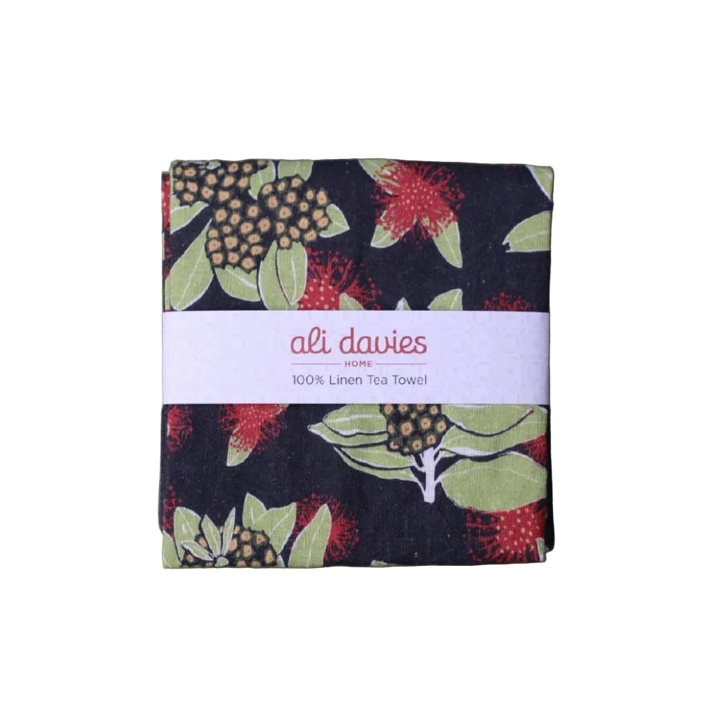 A folded linen tea towel with a botanical print of red flowers and green leaves on a dark navy background, wrapped with a white paper band that reads 'ali davies HOME 100% Linen Tea Towel'. The towel is photographed flat against a white  background.