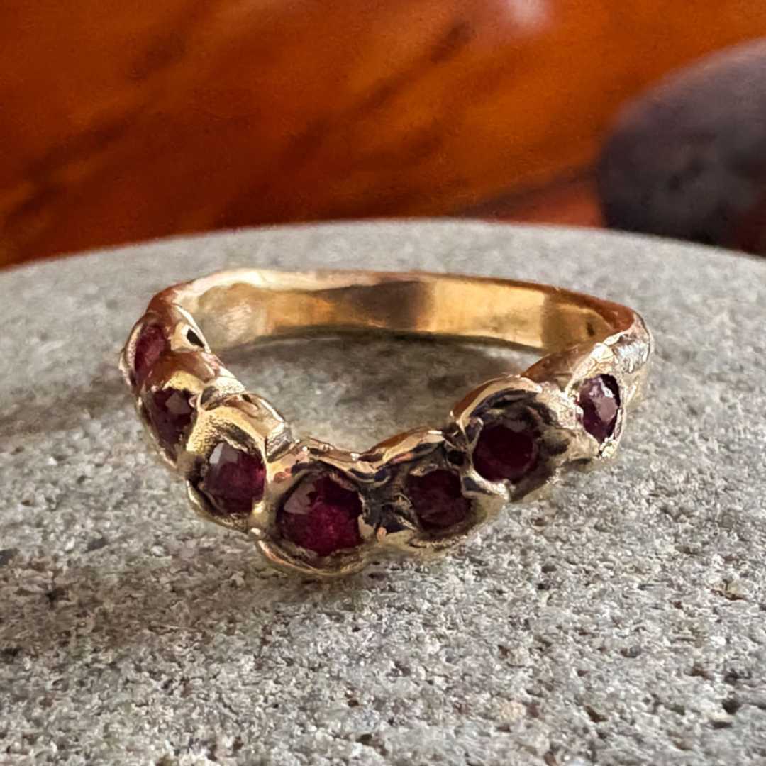 A gold ring featuring a row of round dark red gemstones, resting on a textured gray stone surface with a blurred wooden background.