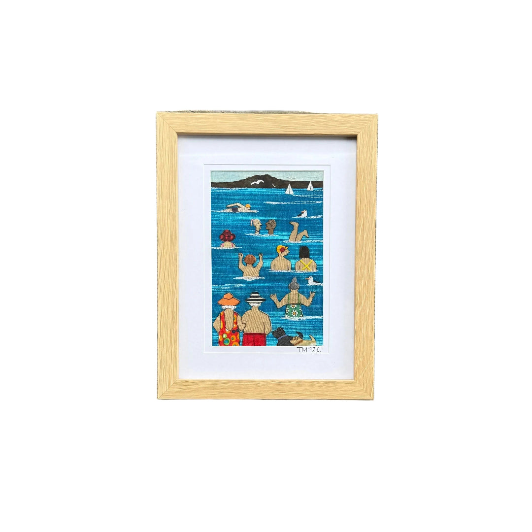 A textile & embroidered artwork depicting a whimsical seaside swimming scene. The artwork features stylized figures in the blue ocean, with a black mountain silhouette and white sailboats in the background.