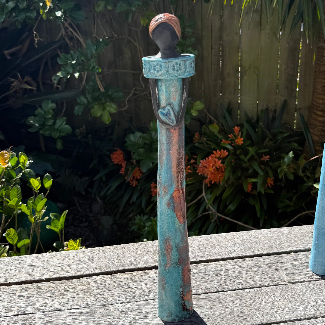 A tall lady ceramic figure with a distressed turquoise-blue and rust-orange glaze, holding a heart-shaped cutout , photographed outdoors on a wooden table with garden foliage and a fence in the background.