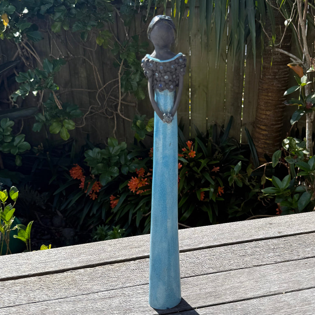 A tall lady ceramic figure with an aqua glazed dress & dark floral bodice, holding a silvery flower, photographed outdoors on a wooden table with garden foliage and a fence in the background.
