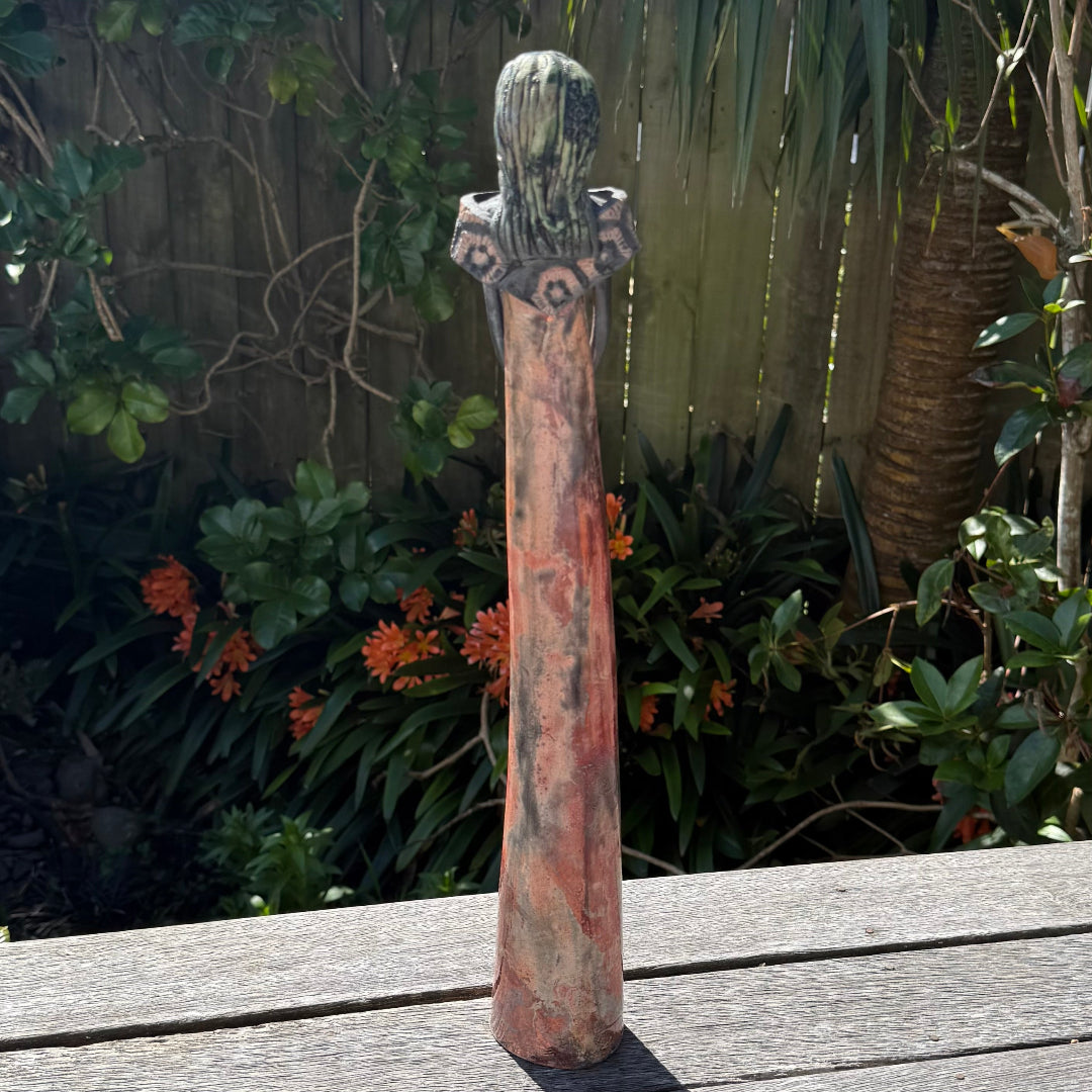 A back view of a tall lady ceramic figure with a shiny copper glazed dress with patterned collar, photographed outdoors on a wooden table with garden foliage and a fence in the background.