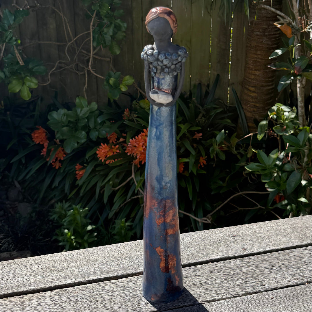 A tall lady ceramic figure with a distressed dark blue and rust-orange glaze, holding a swaddled baby, photographed outdoors on a wooden table with garden foliage and a fence in the background.