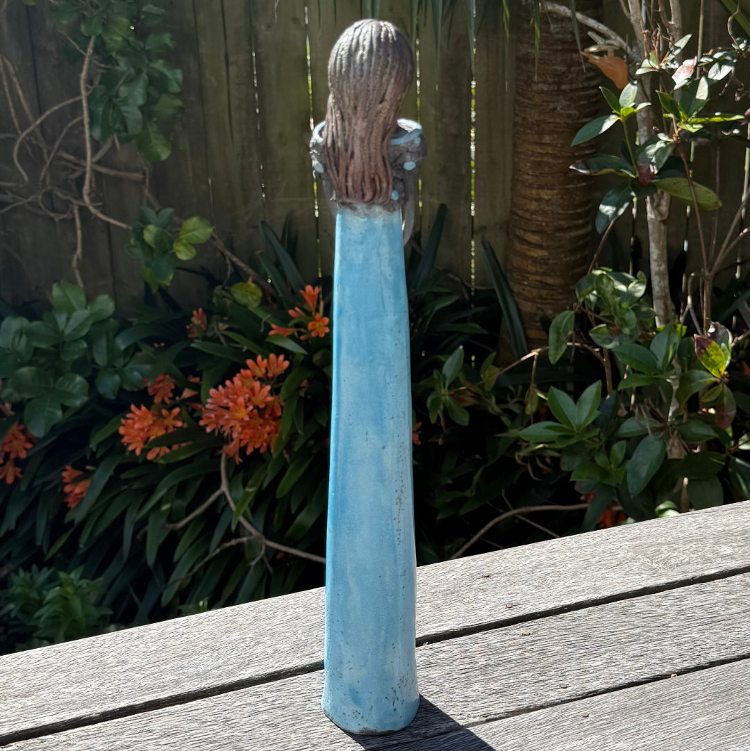 Back view of a tall lady ceramic figure with an aqua glazed dress & dark floral bodice, photographed outdoors on a wooden table with garden foliage and a fence in the background.