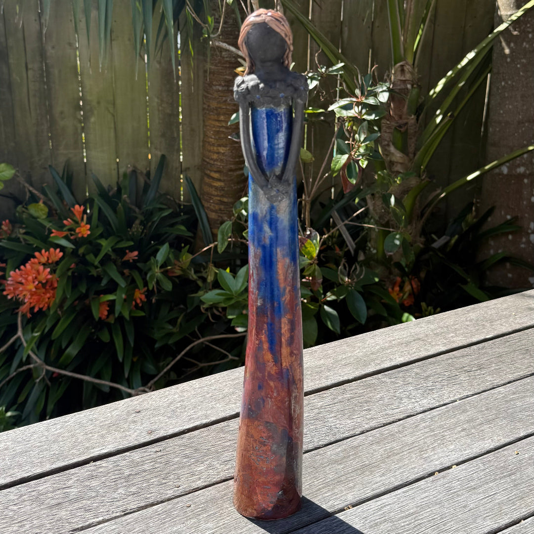 A tall lady ceramic figure with a distressed royal blue and copper glaze, holding a dark flower, photographed outdoors on a wooden table with garden foliage and a fence in the background.