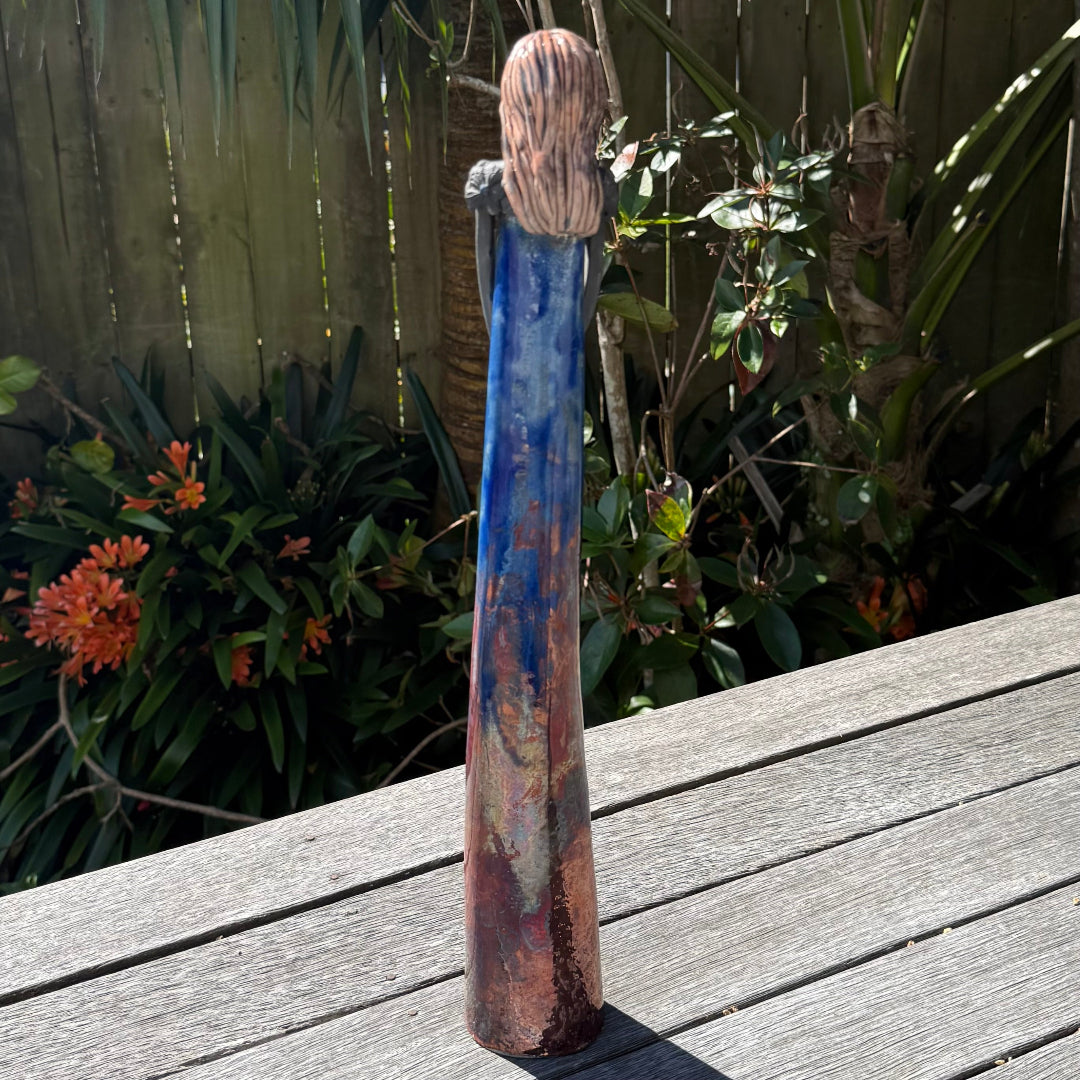 Back view of a tall lady ceramic figure with a distressed royal blue and copper glaze, photographed outdoors on a wooden table with garden foliage and a fence in the background.