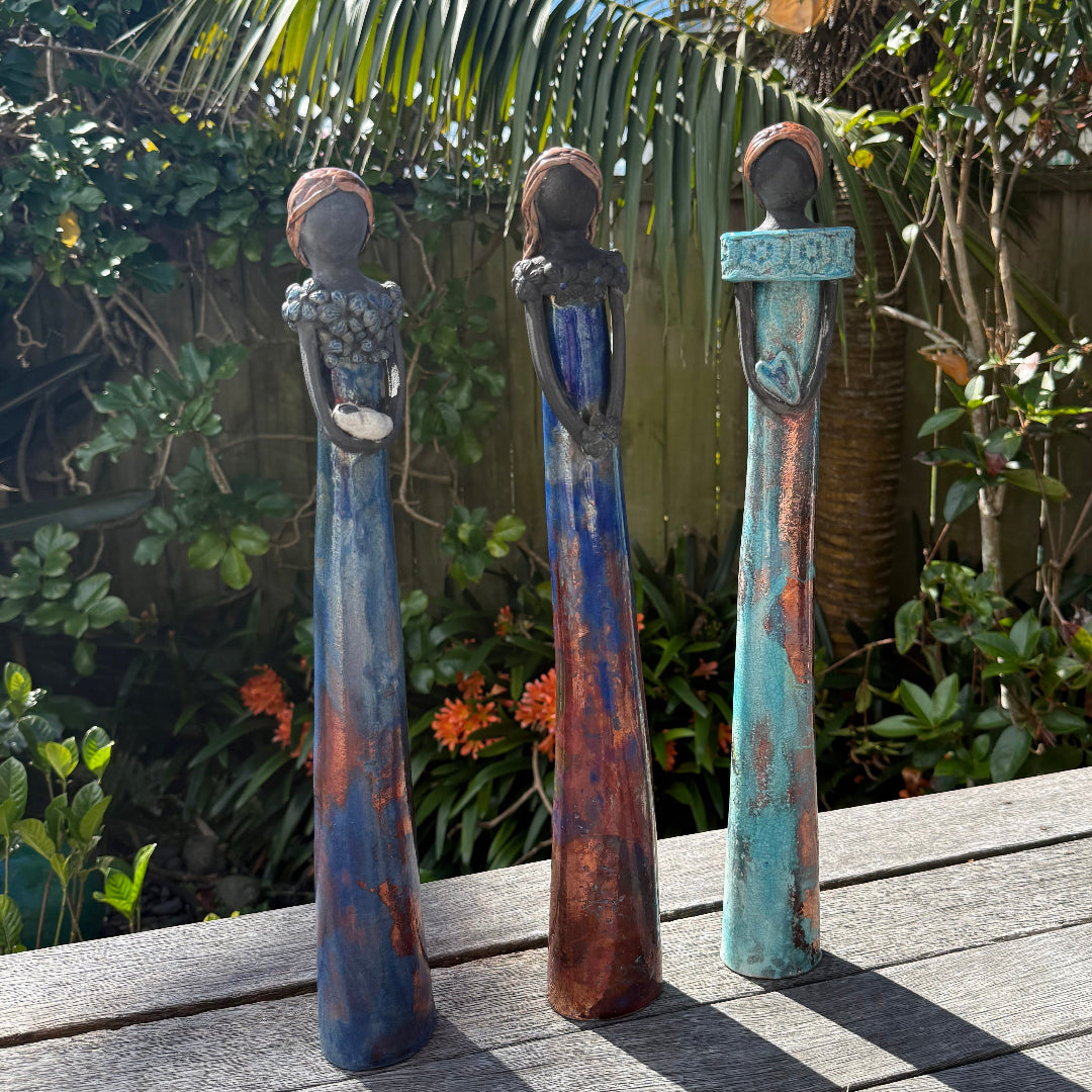 Three tall ceramic ladies figurines with black faces and elongated bodies stand on a weathered wooden deck outdoors, with tropical plants and a blue sky in the background.