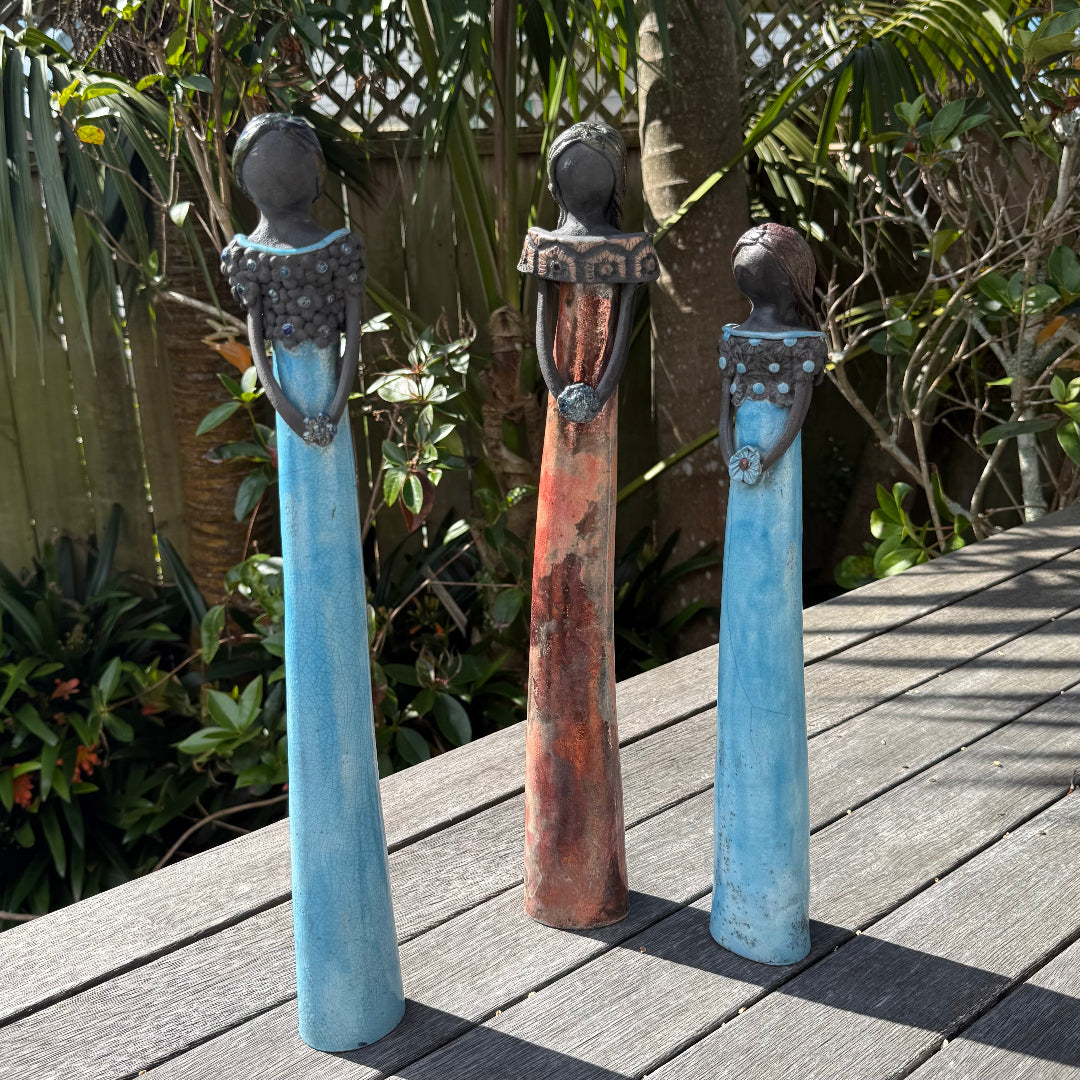 Three tall ceramic ladies figurines with black faces and elongated bodies stand on a weathered wooden deck outdoors, with tropical plants and a blue sky in the background.