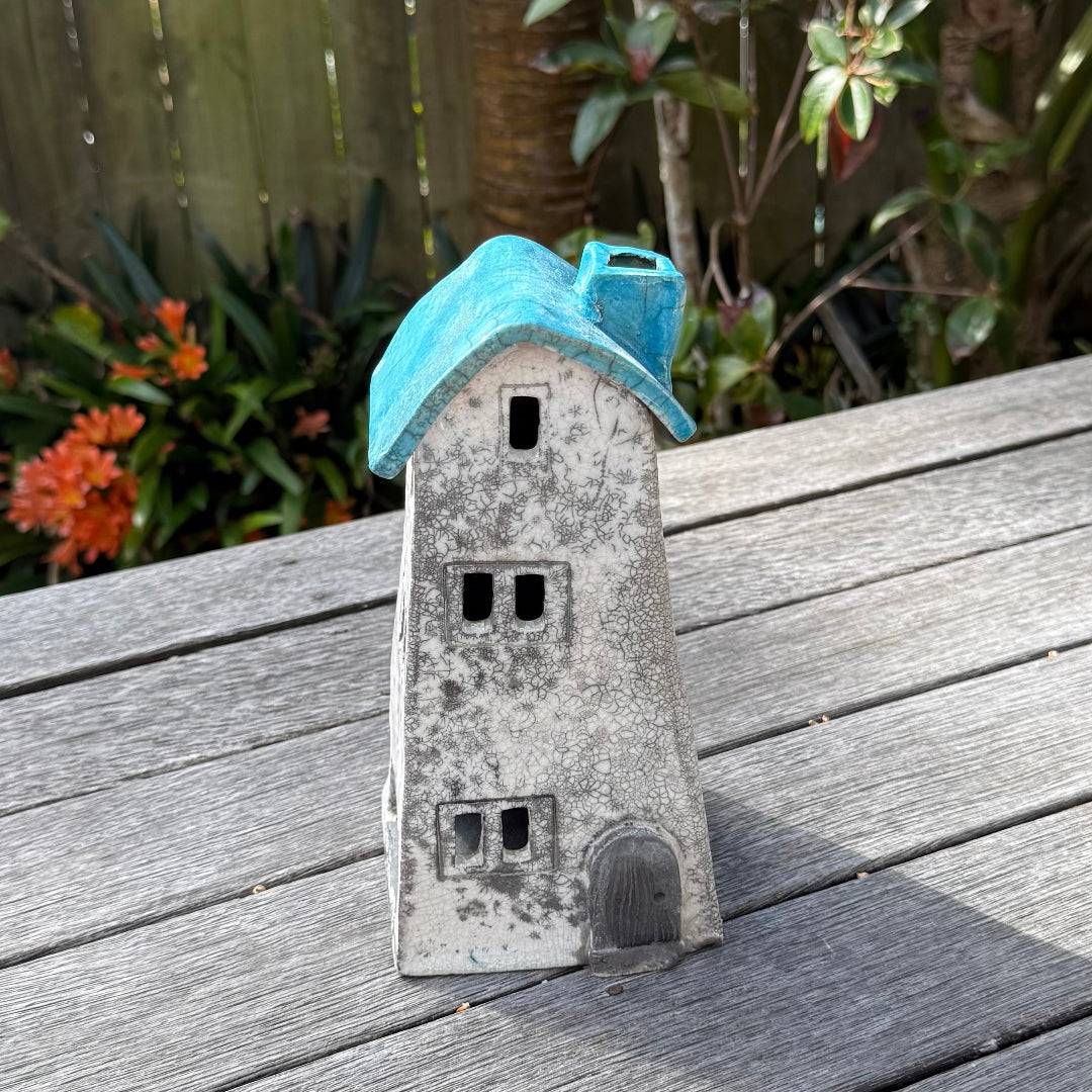 A ceramic house-shaped tealight holder  with a distressed white body, dark gray window cutouts, and an aqua roof, photographed outdoors on a weathered wooden surface with garden plants in the background.