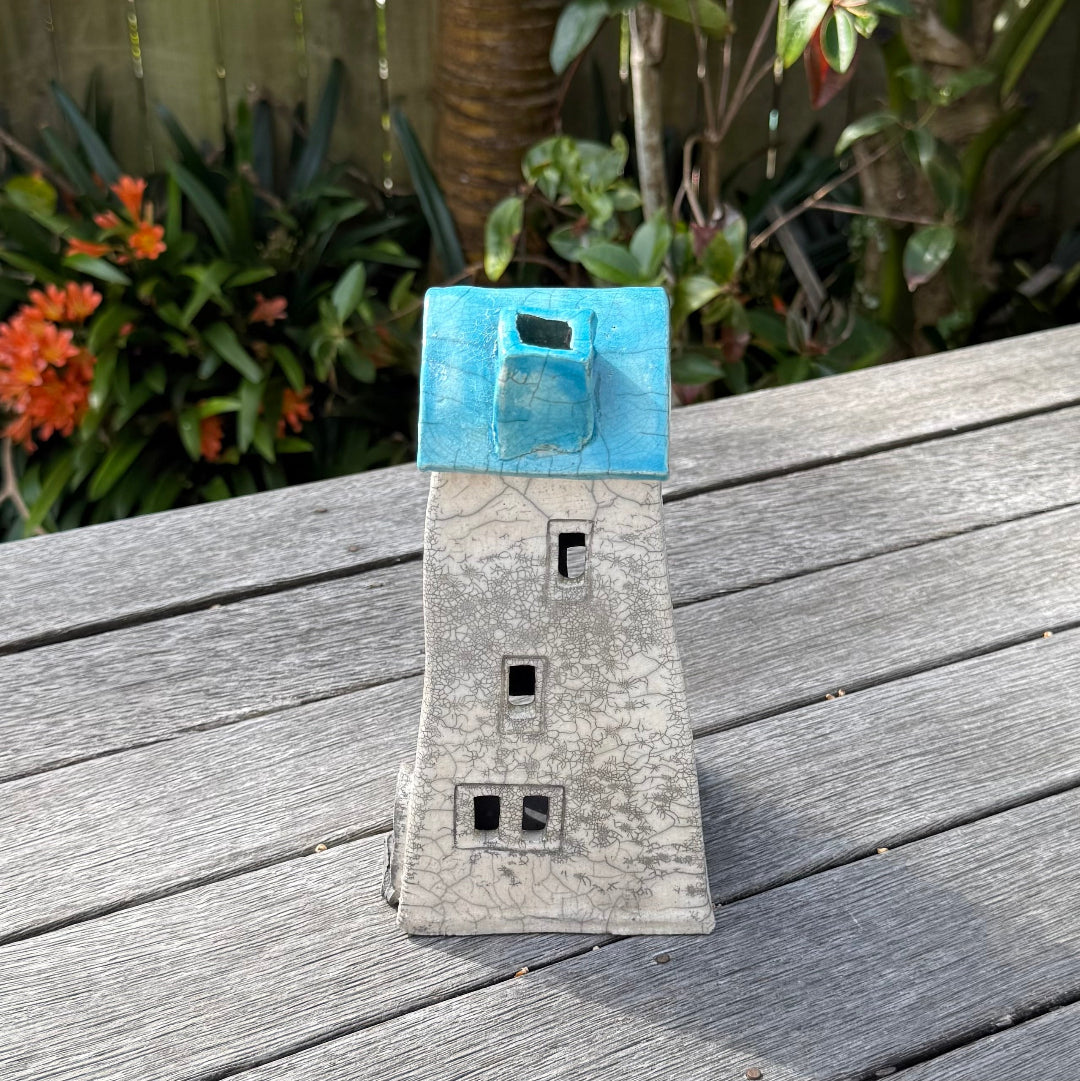 A ceramic house-shaped tealight holder  with a distressed white body, dark gray window cutouts, and an aqua roof, photographed outdoors on a weathered wooden surface with garden plants in the background.