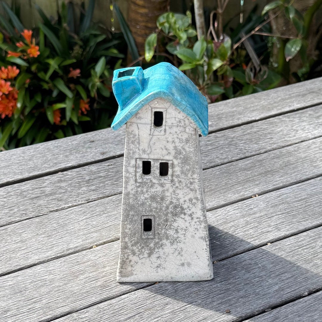 A ceramic house-shaped tealight holder  with a distressed white body, dark gray window cutouts, and an aqua roof, photographed outdoors on a weathered wooden surface with garden plants in the background.