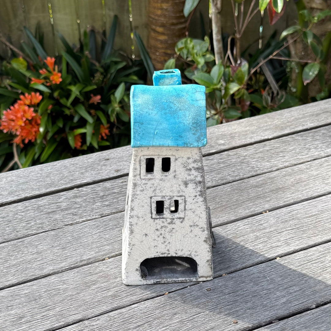 A ceramic house-shaped tealight holder  with a distressed white body, dark gray window cutouts, and an aqua roof, photographed outdoors on a weathered wooden surface with garden plants in the background.