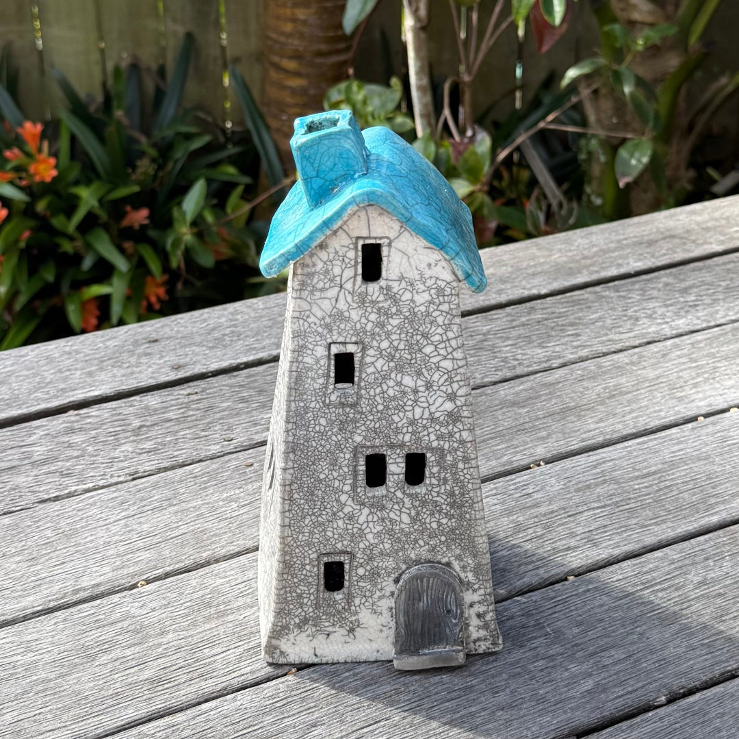A ceramic house-shaped tealight holder  with a distressed white body, dark gray window cutouts, and an aqua roof, photographed outdoors on a weathered wooden surface with garden plants in the background.