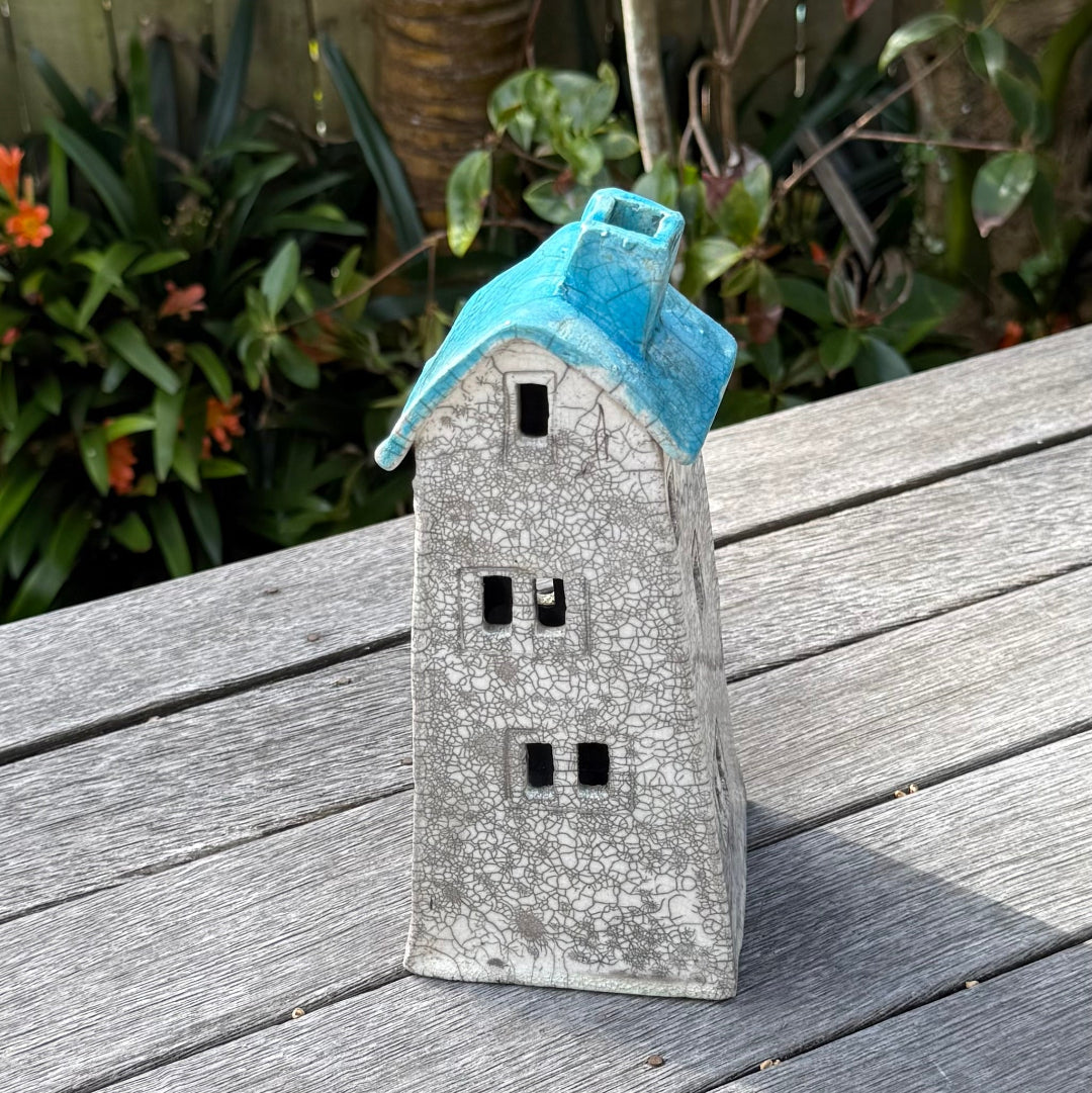A ceramic house-shaped tealight holder  with a distressed white body, dark gray window cutouts, and an aqua roof, photographed outdoors on a weathered wooden surface with garden plants in the background.