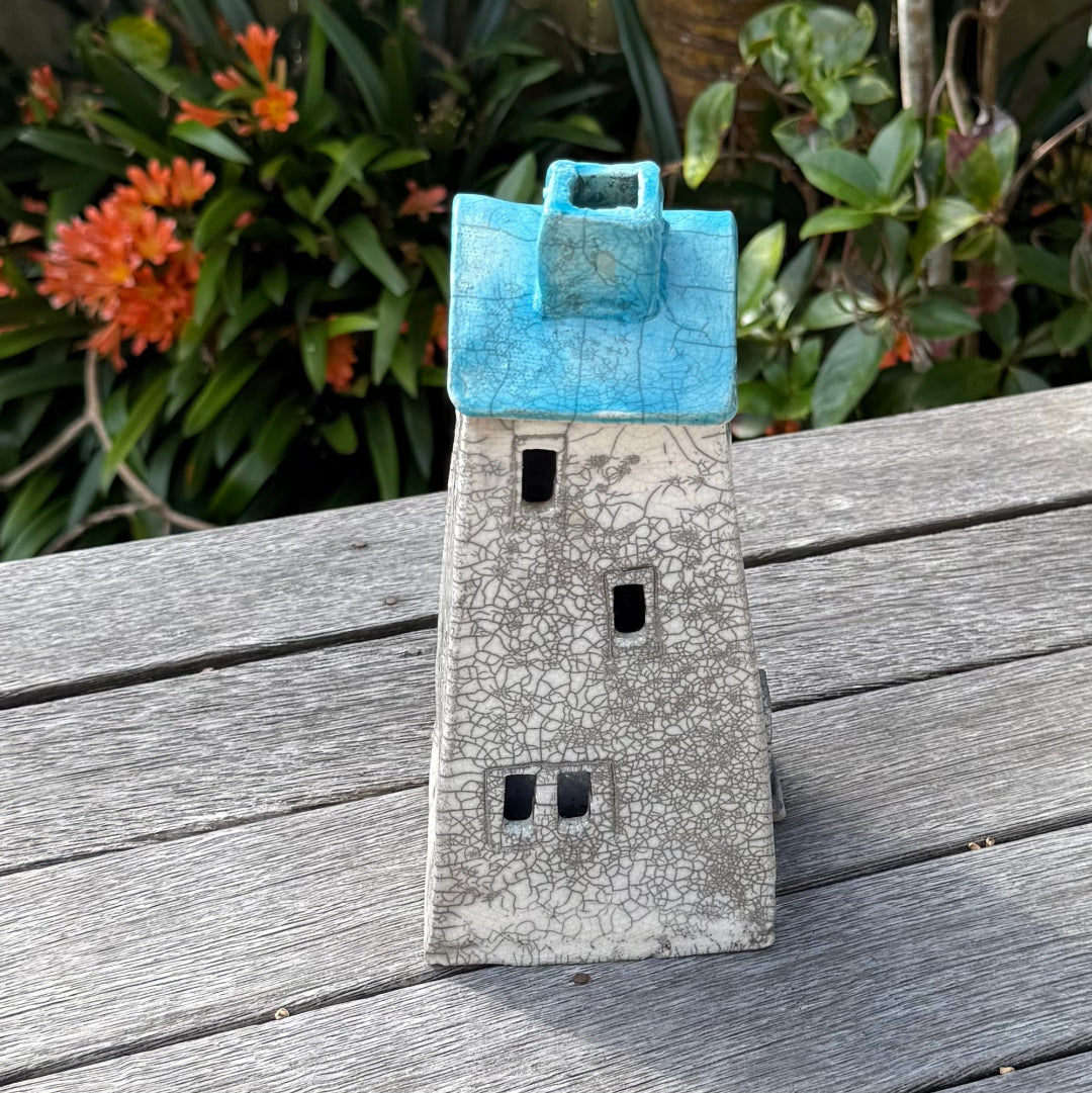 A ceramic house-shaped tealight holder  with a distressed white body, dark gray window cutouts, and an aqua roof, photographed outdoors on a weathered wooden surface with garden plants in the background.