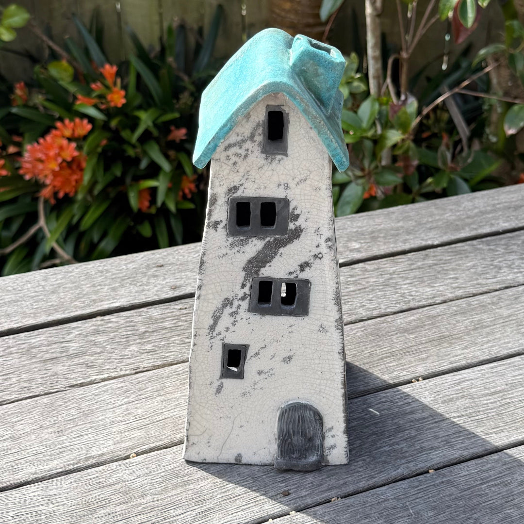 A ceramic house-shaped tealight holder  with a distressed white body, dark gray window cutouts, and a turquoise roof, photographed outdoors on a weathered wooden surface with garden plants in the background.
