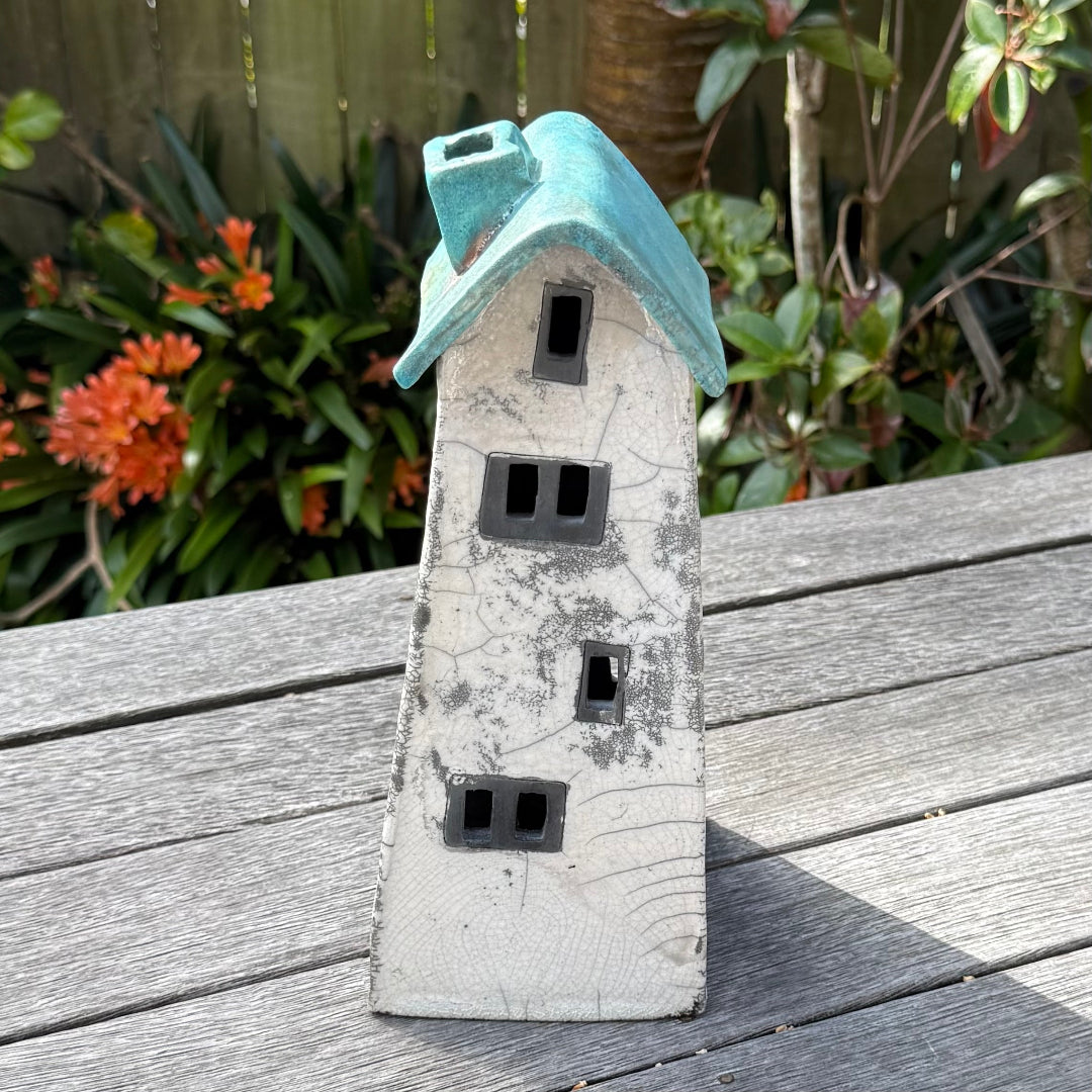 A ceramic house-shaped tealight holder  with a distressed white body, dark gray window cutouts, and a turquoise roof, photographed outdoors on a weathered wooden surface with garden plants in the background.