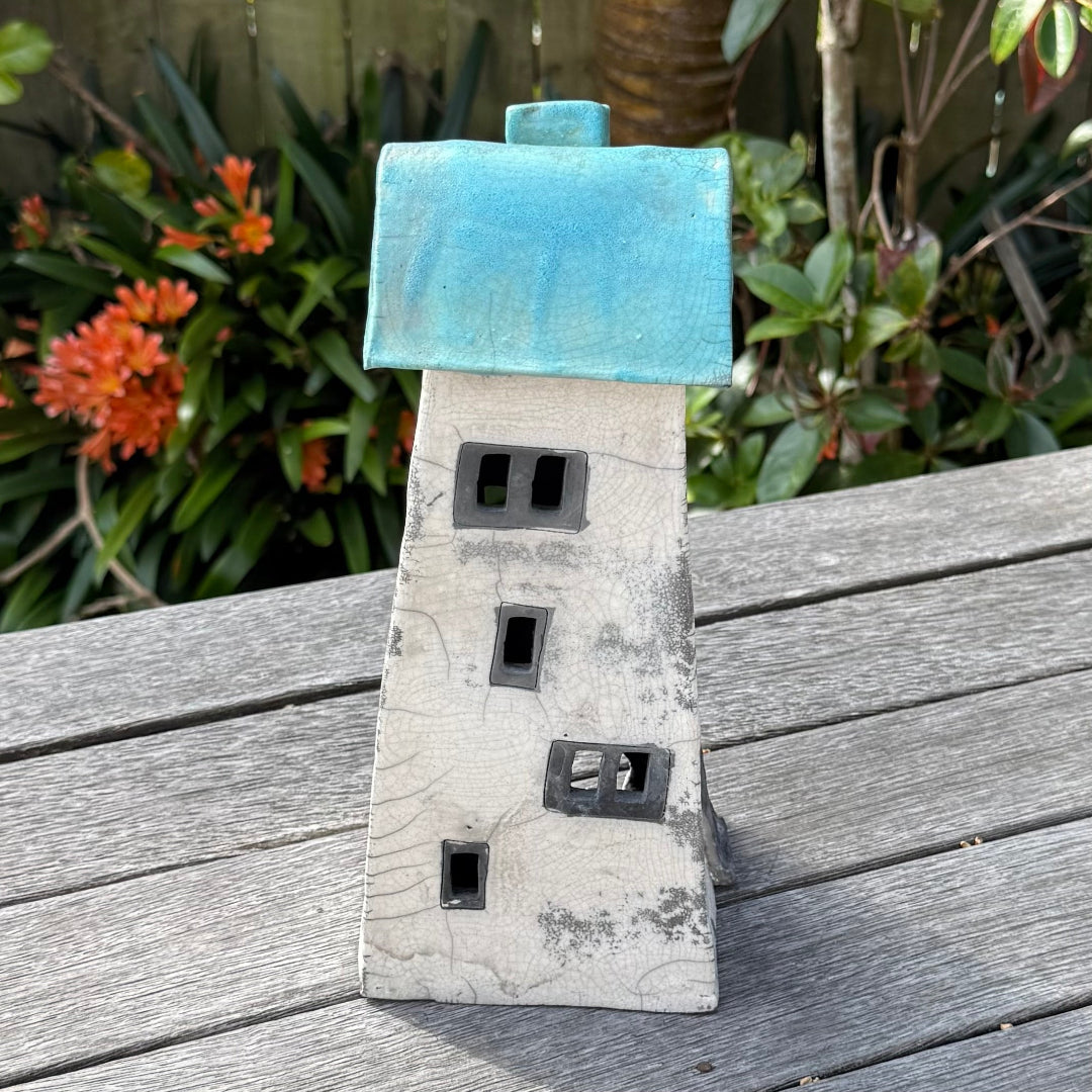 A ceramic house-shaped tealight holder  with a distressed white body, dark gray window cutouts, and a turquoise roof, photographed outdoors on a weathered wooden surface with garden plants in the background.