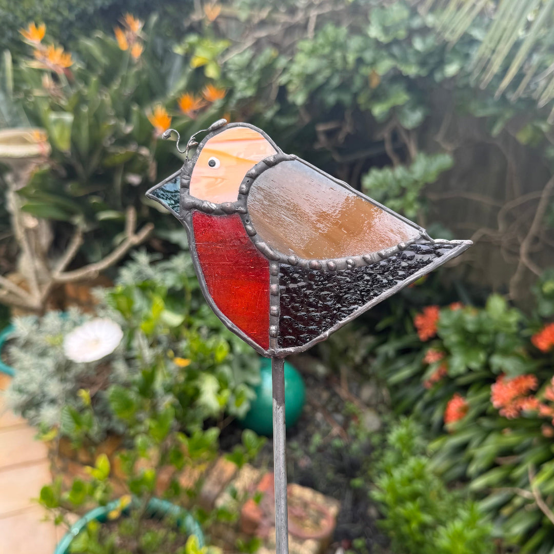 A stained glass bird garden stake featuring a robin design with red, brown, black, and orange glass panels, set against a blurred garden background.