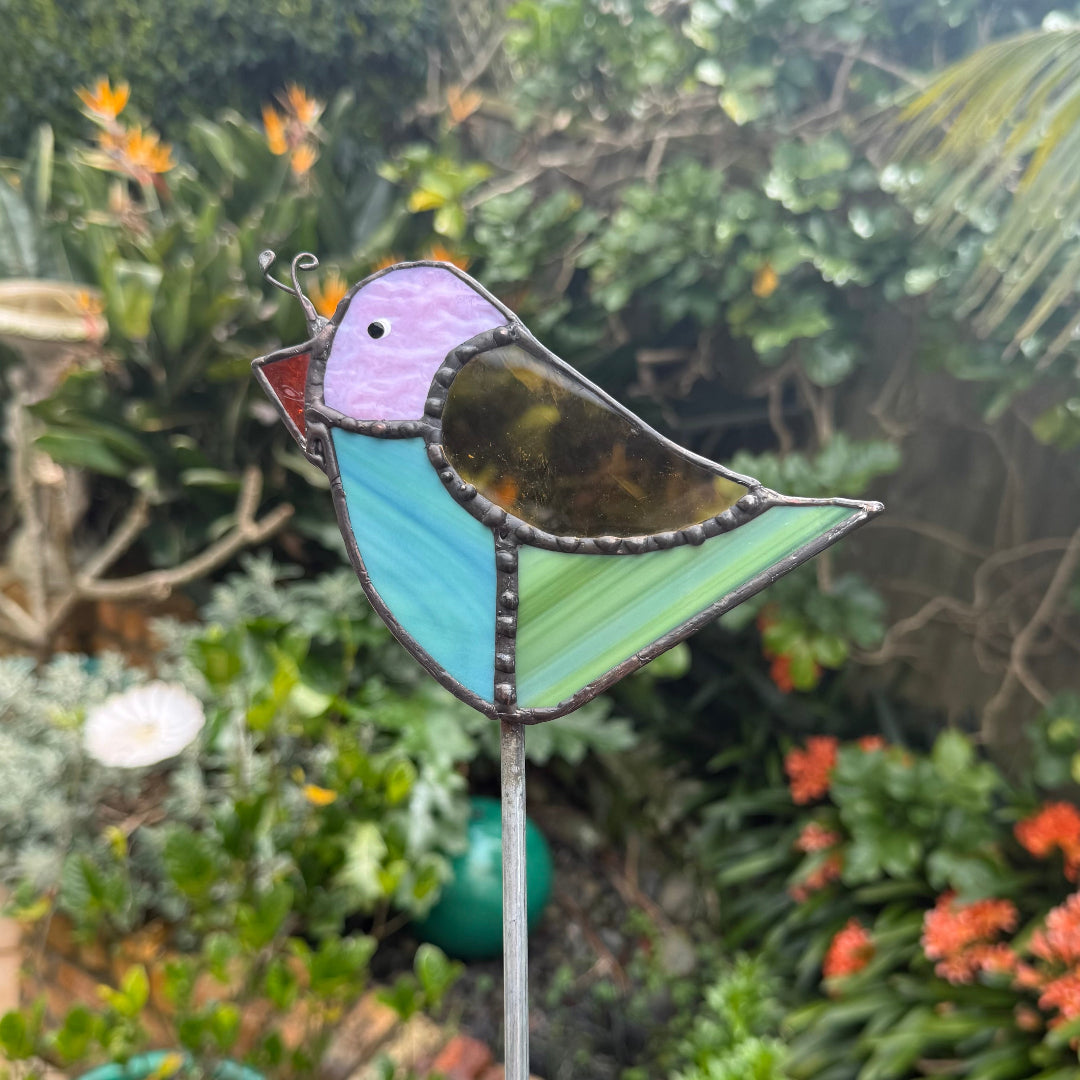 A stained glass bird garden stake featuring a robin design with purple, red, brown, blue and green glass panels, set against a blurred garden background.