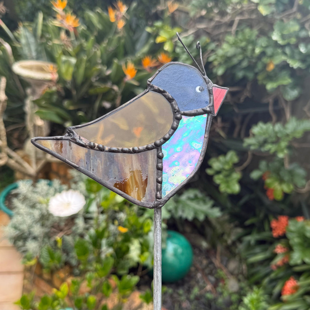 A stained glass bird garden stake featuring a robin design with royal, red, brown, black, and orange glass panels, set against a blurred garden background.