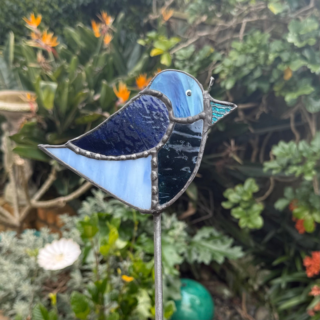 A stained glass bird garden stake featuring shades of blue, navy, and light blue, set against a blurred garden background with green foliage and orange flowers.