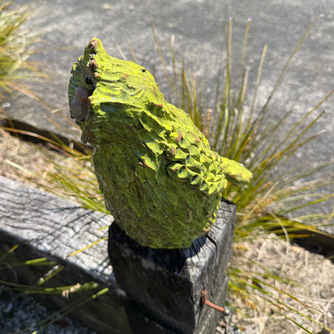 Side view of a green standing pottery Kakapo bird on a post in a garden.