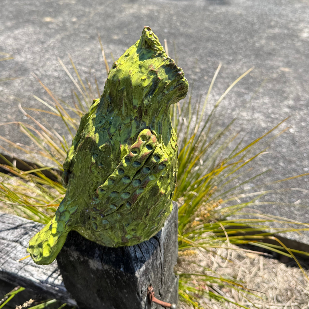 Side view of a green standing pottery Kakapo bird on a post in a garden.