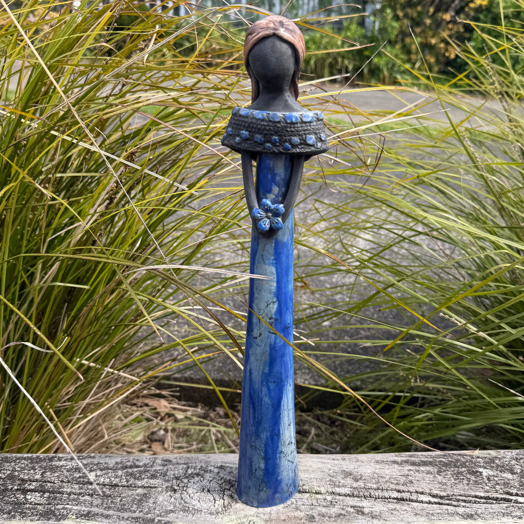 A decorative figurine of a woman with light hair, wearing a long blue dress and holding a blue flower, standing on a weathered wooden table with a garden background.