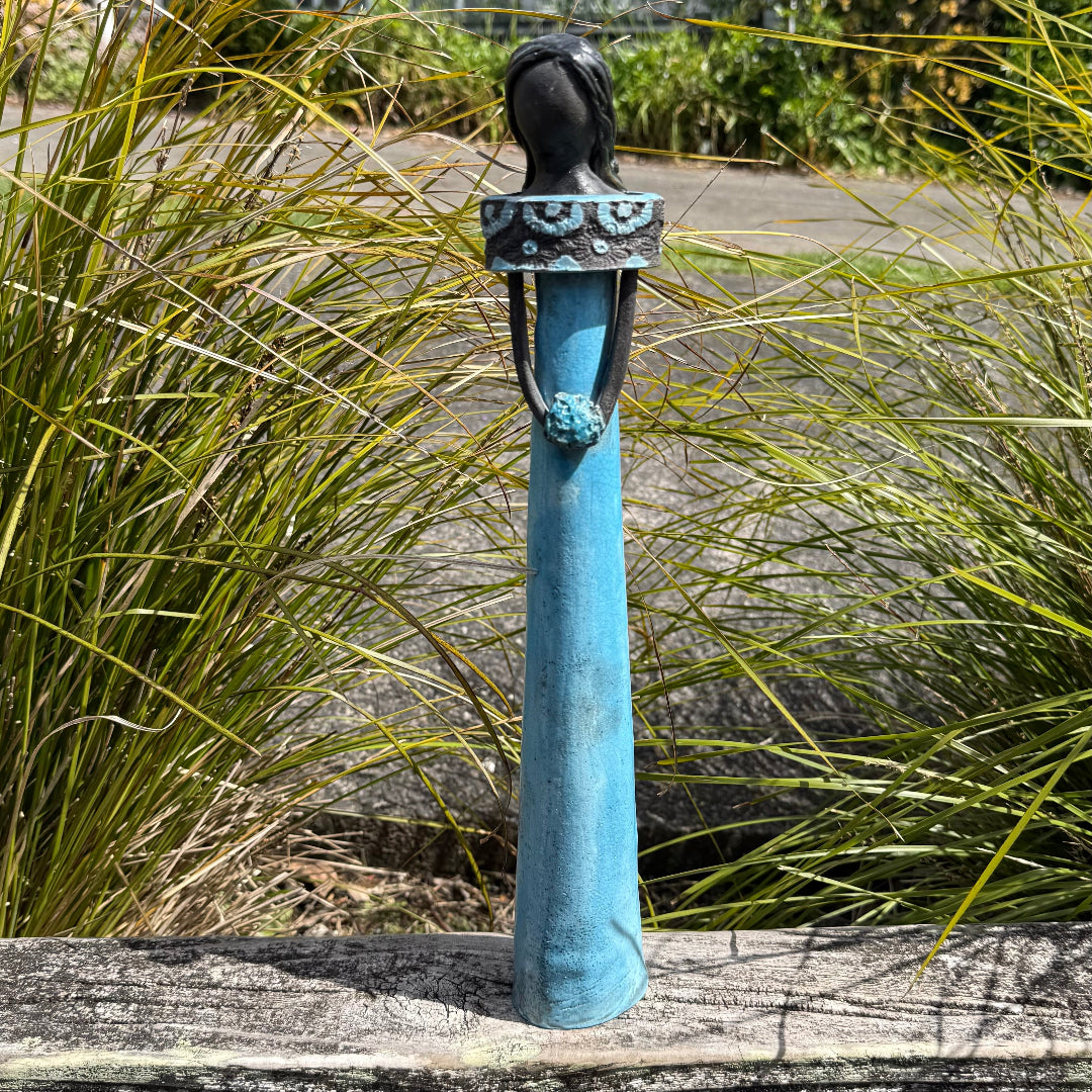 A ceramic decorative figurine of a woman with dark hair, wearing a long aqua dress and holding an aqua posy, standing on a weathered wooden table with a garden background.