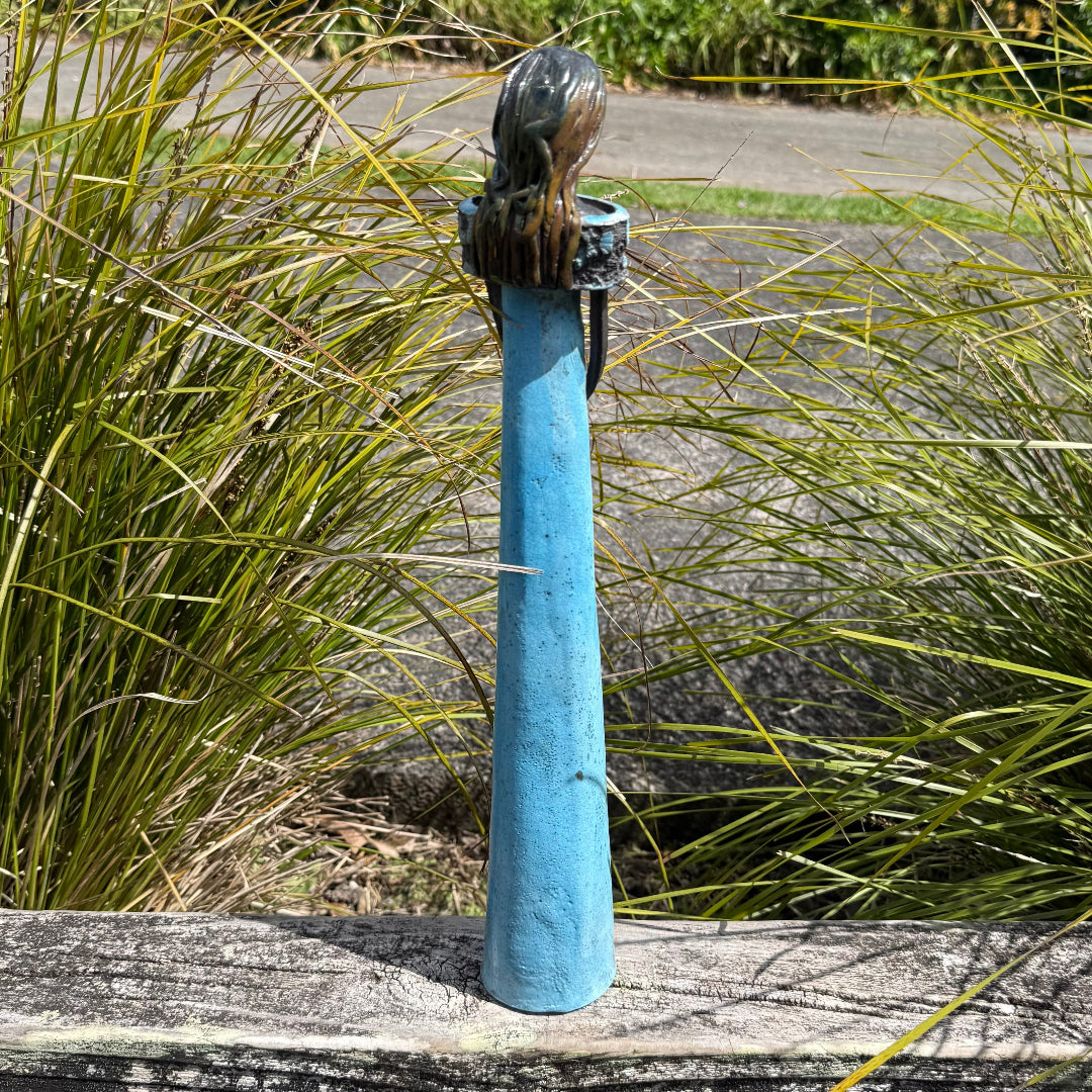 Back view of a ceramic decorative statue of a woman with long darkish hair and an aqua dress, standing on a weathered wooden deck with garden foliage in the background.