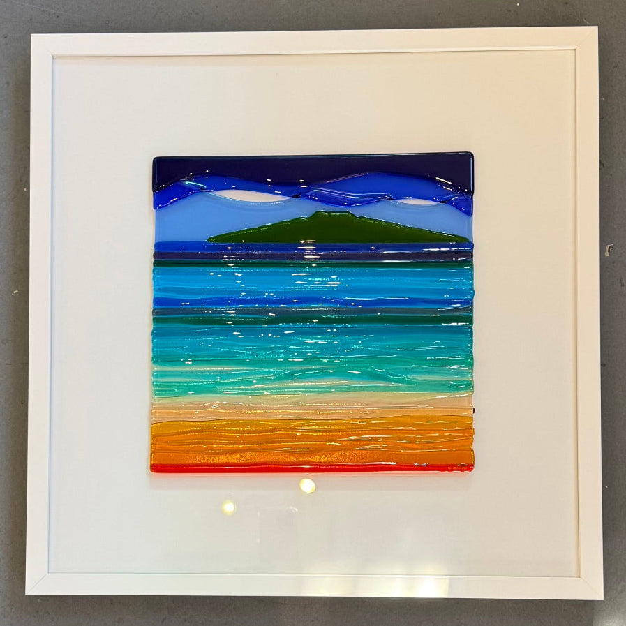 A square framed artwork depicting a colorful beach or ocean scene with blue sky, green land, turquoise water, and orange sand, mounted on a gray wall.