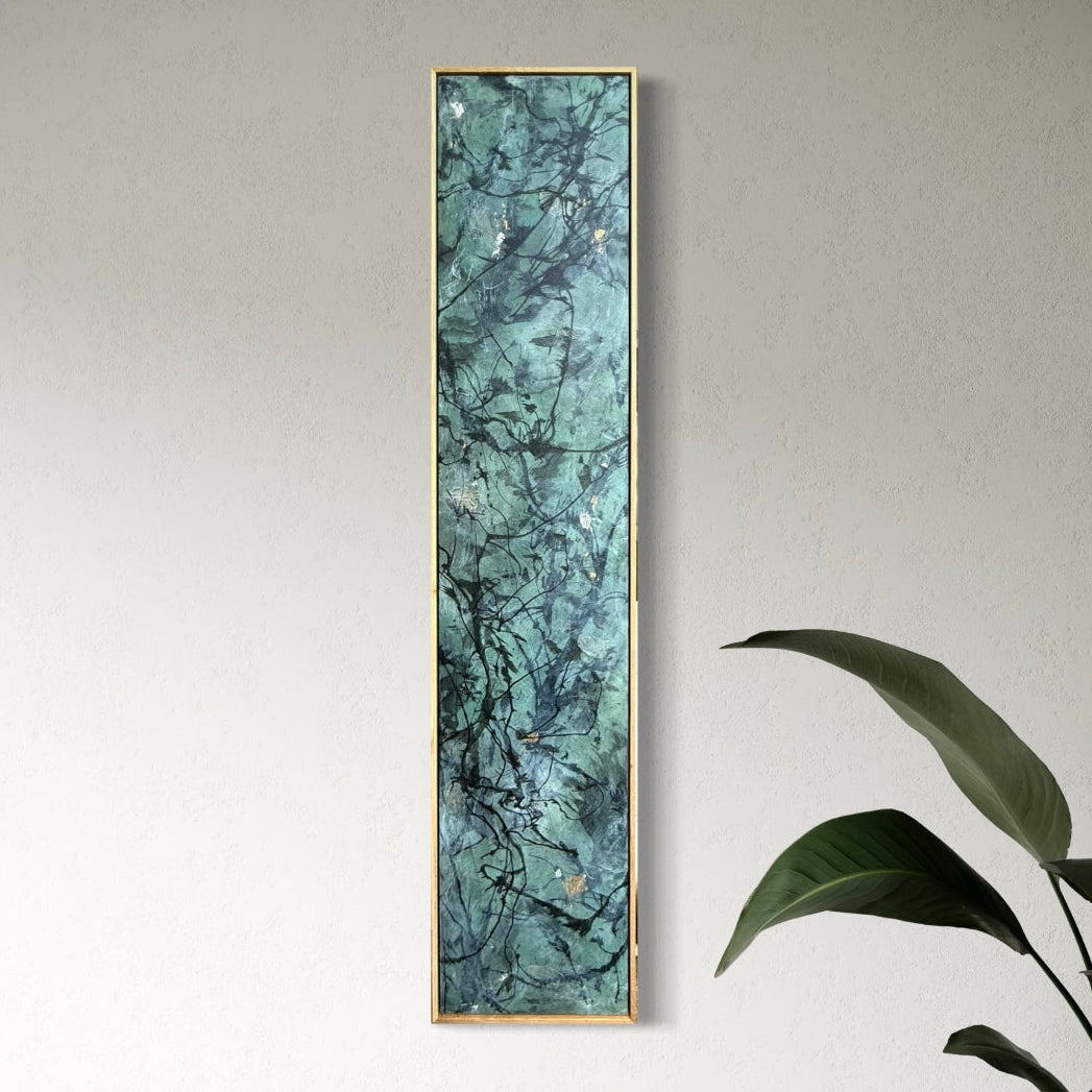 A tall, narrow framed wall art piece with a teal and turquoise marble pattern and a thin gold frame, hanging on a light gray wall in a styled room with a woven chair and a potted plant.