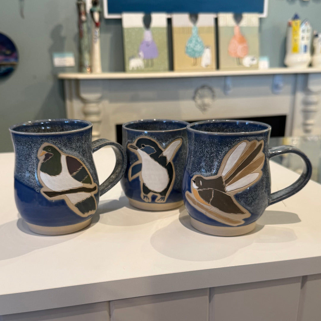 Three ceramic mugs with bird designs on a white surface, with a blurred background of shelves and items.