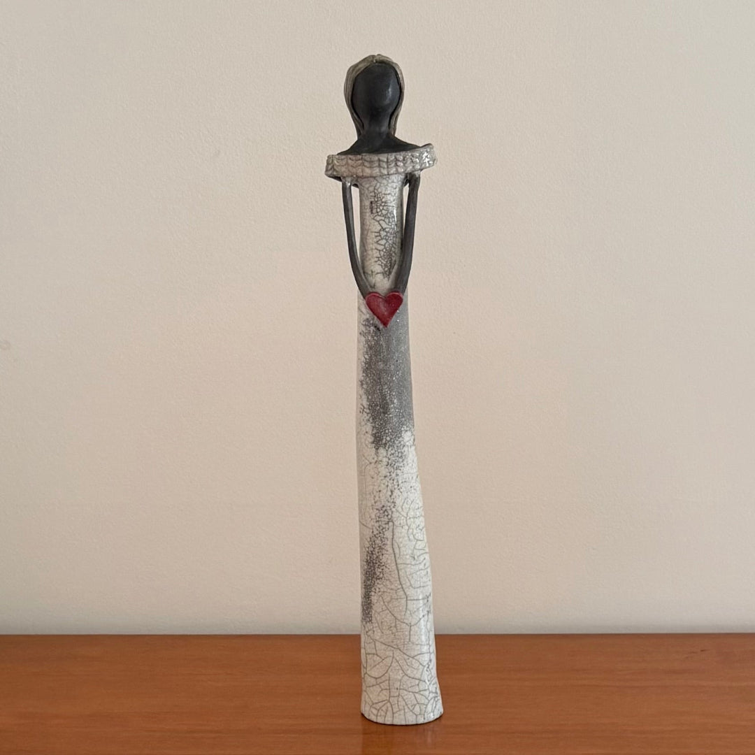 Ceramic sculpture of a figure with a red heart on a wooden surface and beige wall background