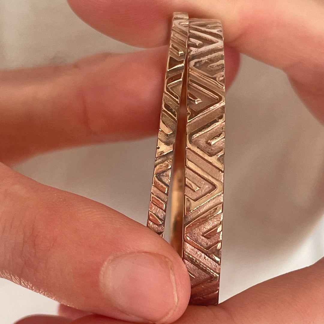 Copper & silver bangles with geometric pattern held between fingers against a neutral background