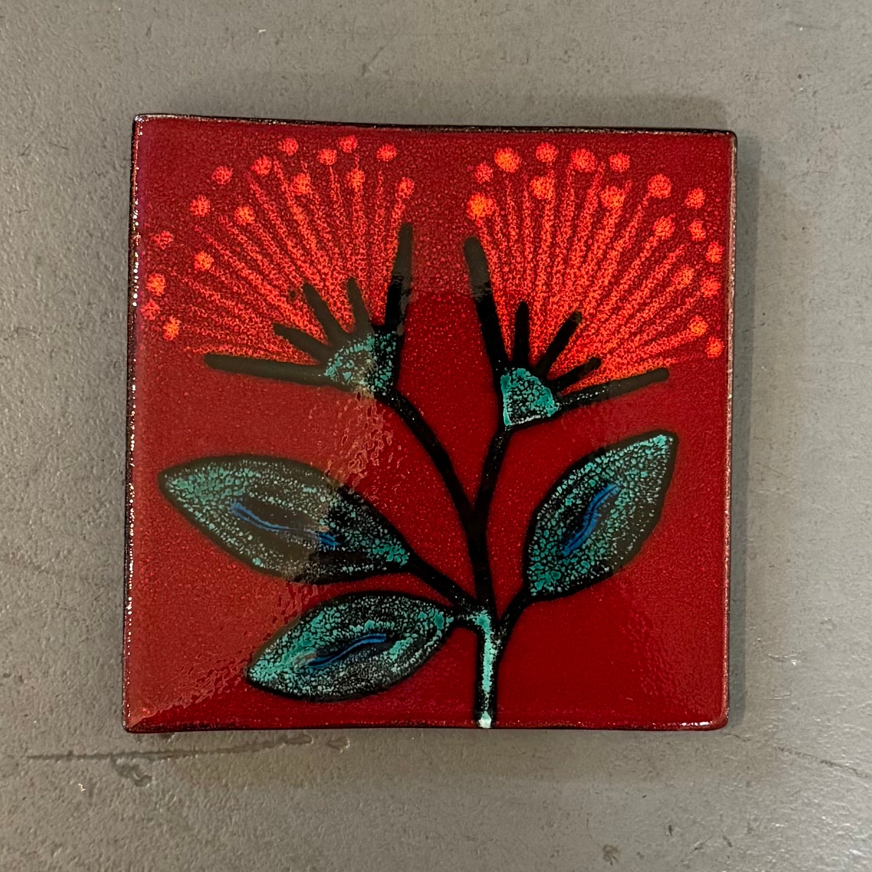 Square red tile with pohutakawa fower design on a gray surface