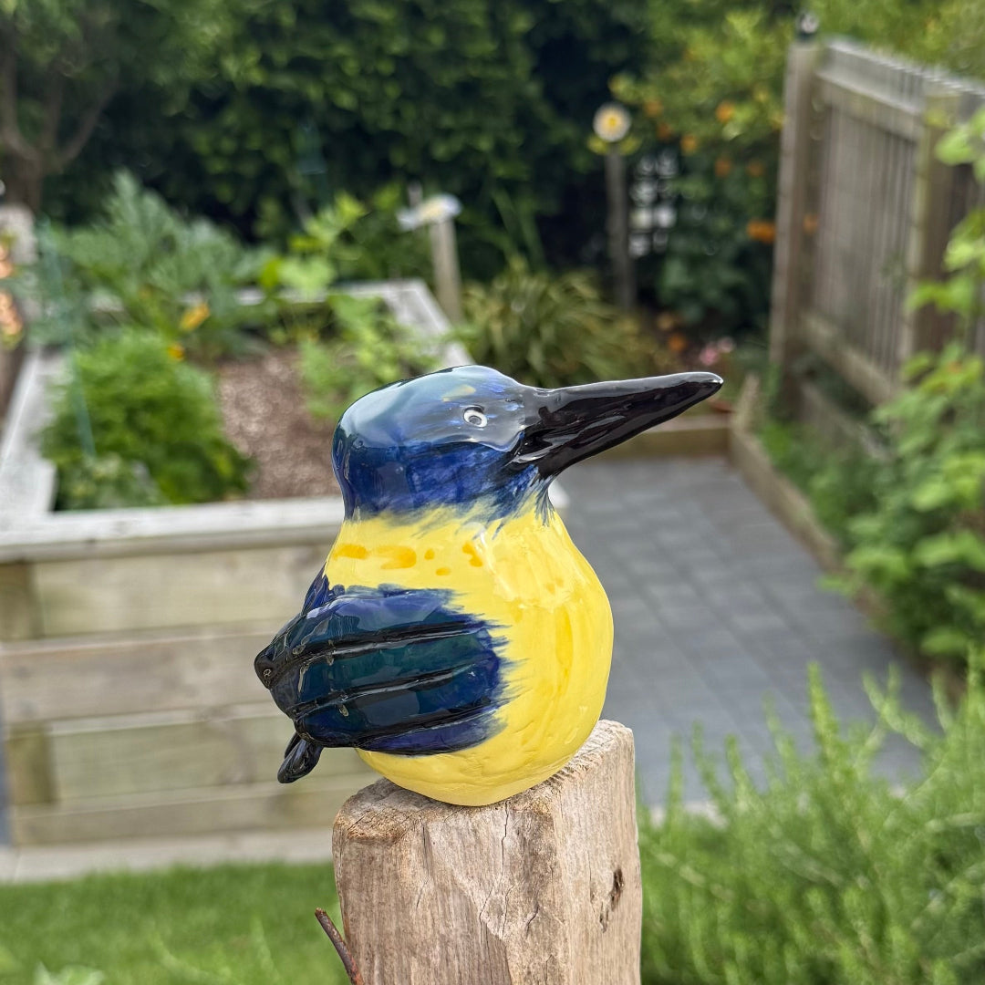 Ceramic Kingfisher bird with blue and yellow colors on a wooden post in a garden setting.