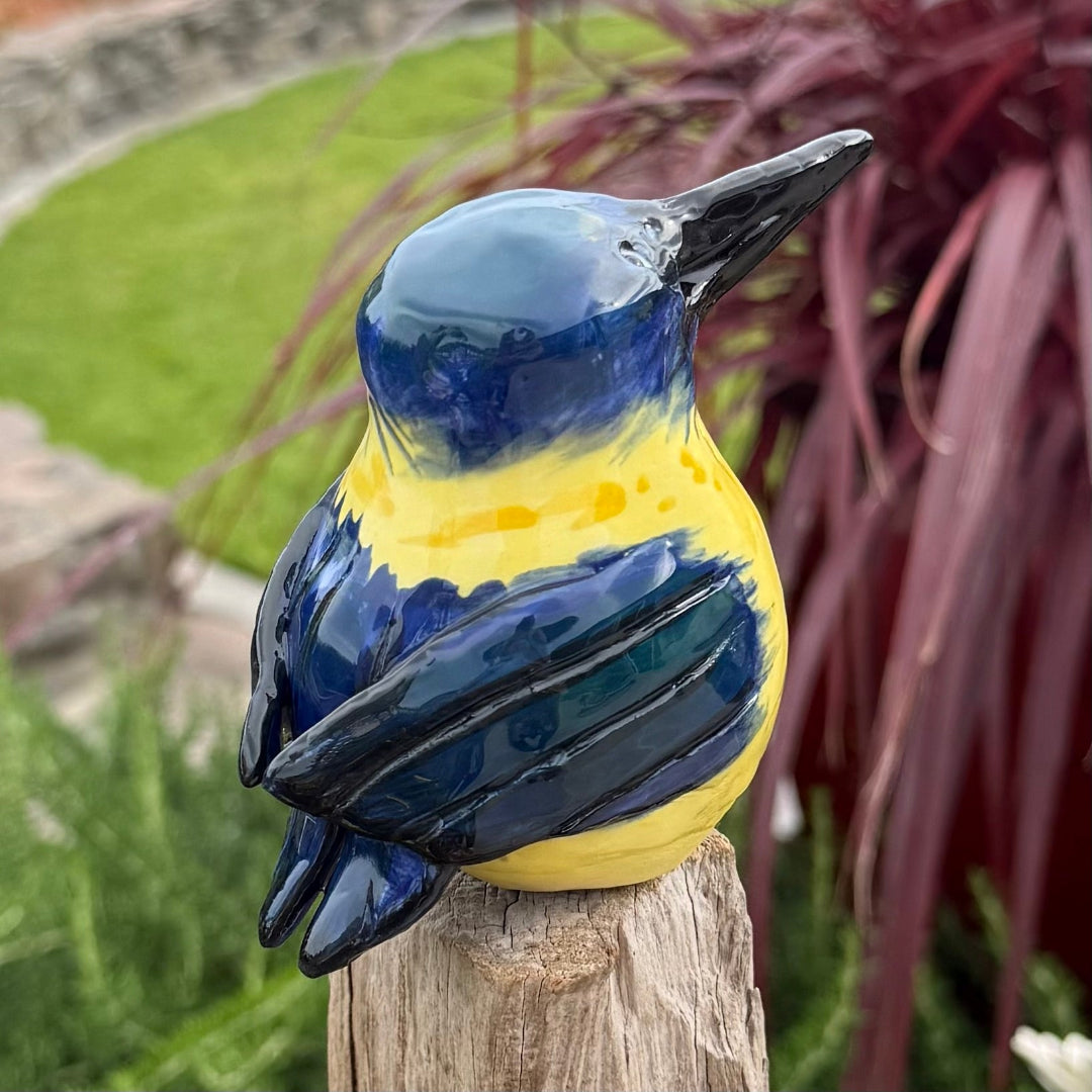 Ceramic Kingfisher bird with blue and yellow colors on a wooden post in a garden setting.