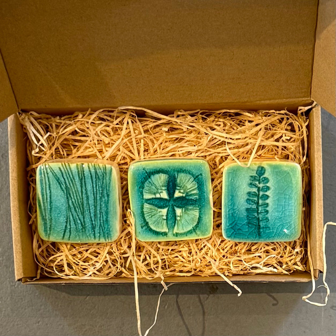 Three green ceramic tiles with plant  designs in a box lined with straw