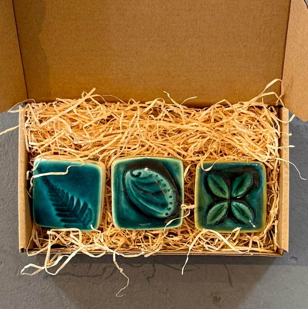 Three green ceramic square tiles in a cardboard box with straw padding.